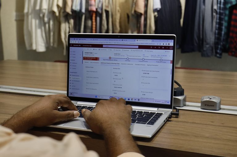 Photo of a person working on a laptop on a desk with a rack of shirts hanging in the background