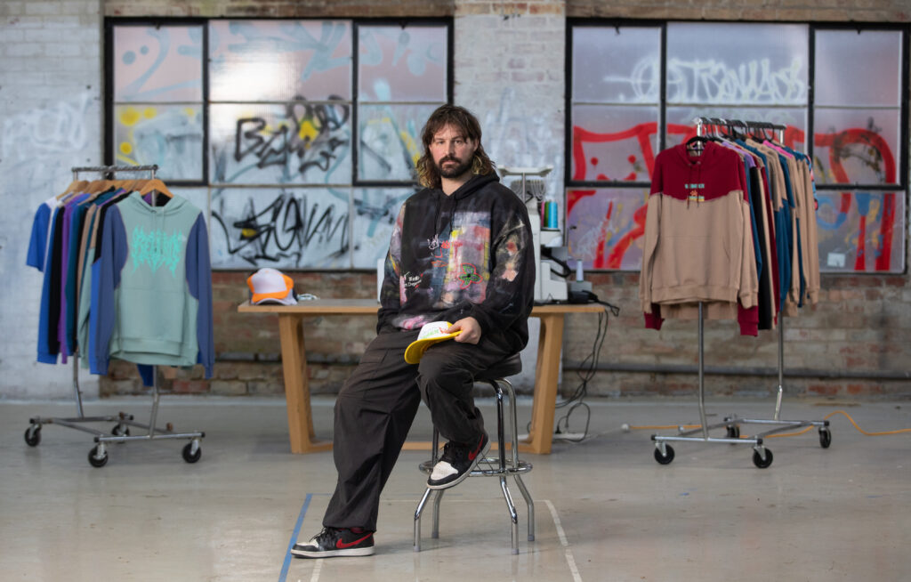 A man with long hair and a beard, sitting on a chair in what seems like a studio or a store with clothes hanging on racks behind him