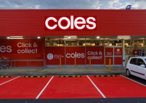 A Coles supermarket storefront with a bright red facade and illuminated 'coles' signage. The store features large windows displaying the interior, and signs for 'Click & Collect' services. A white car is parked in front of the store.