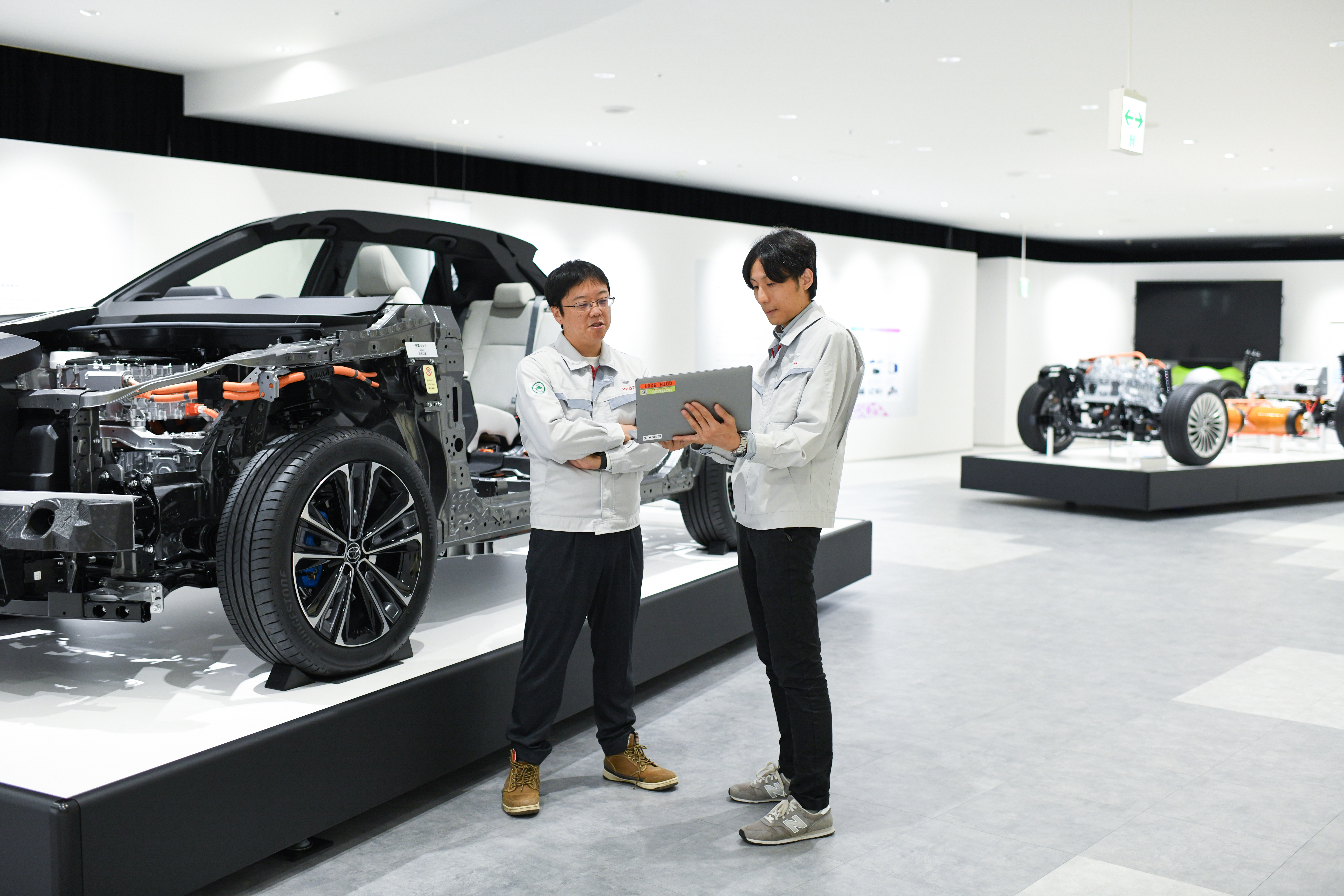 Two men look at a laptop next to a car display with the vehicle body peeled off to show the engine and other internal components.