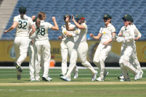 A group of female cricket players in white uniforms celebrate on the field